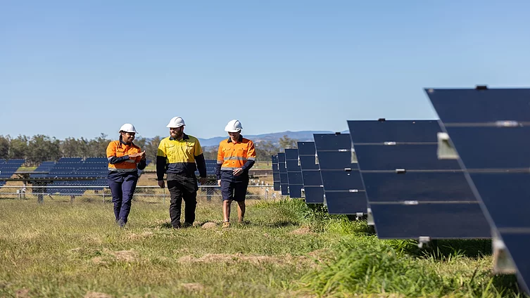 Three technicians in safety helmets and high-visibility workwear walking beside rows of solar panels at a solar farm